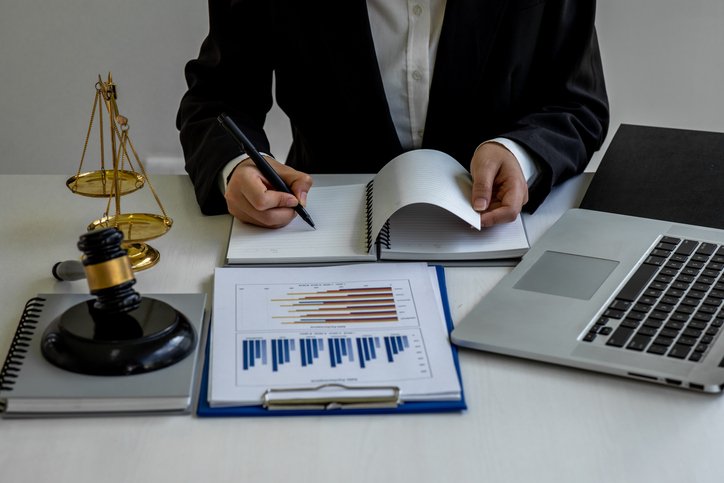lawyer Lawyer at a desk reviewing documents with charts, a notebook, laptop, gavel, and scales of justice nearby.