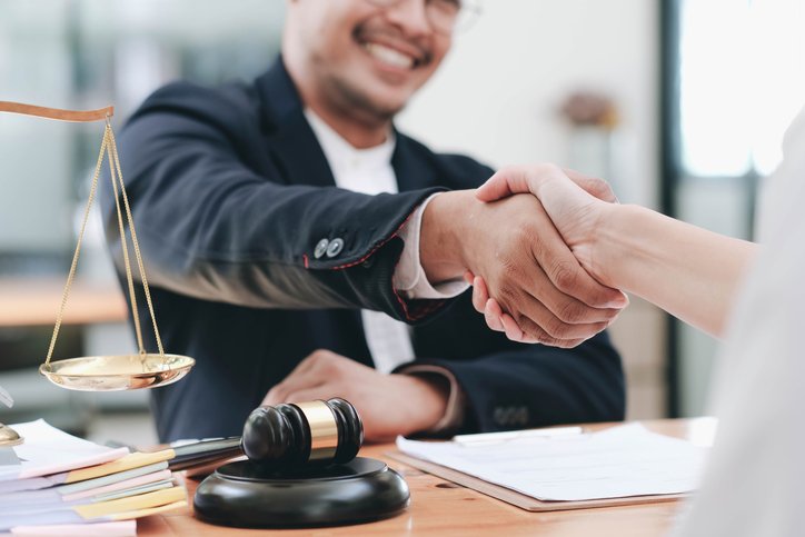 Businessman shaking hands to seal a deal with his partner lawyers or attorneys discussing a contract agreement. Two professionals shake hands across a desk in a legal office, with a gavel and scales of justice nearby.