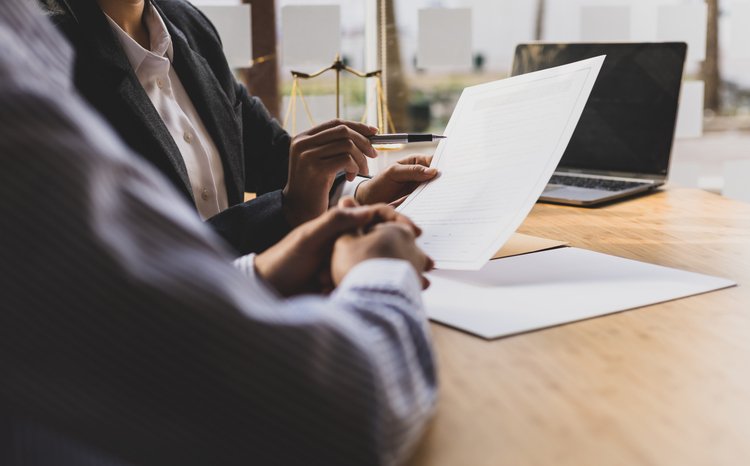 Female lawyer is explaining the terms of the legal contract document and asking the client to sign it properly. Two professionals review a document at a wooden table, with a laptop and scales of justice in the background.