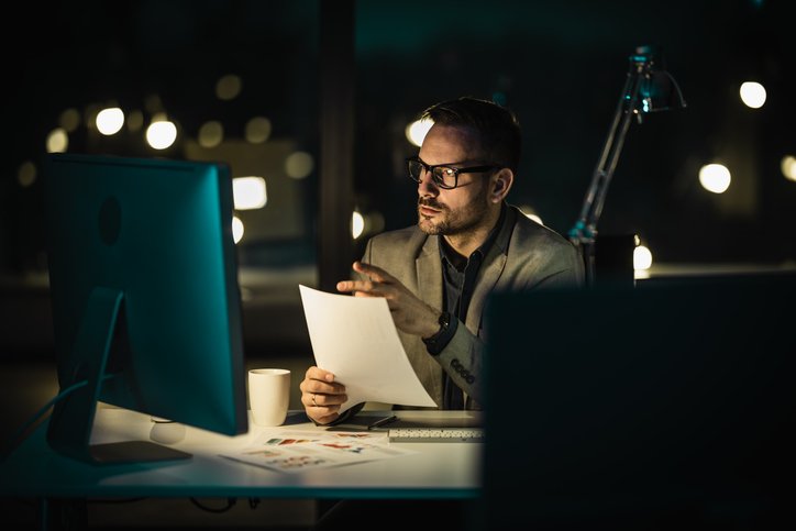 Late-Night Broker-Exhausted but Determined Professional man with glasses reviews documents at a desk with a computer, in a dim, city-lit office at night.