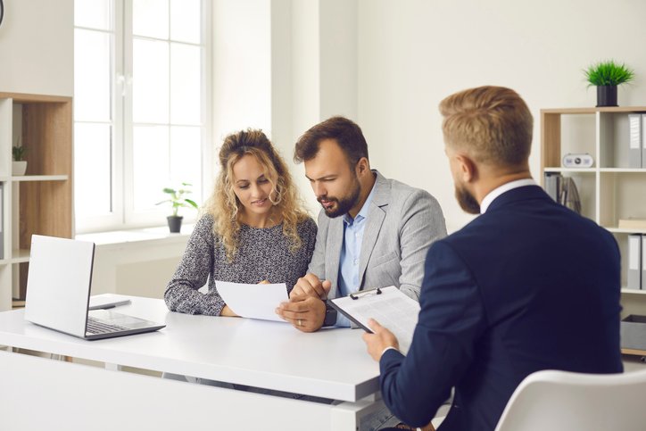Young couple reading some documents sitting at desk in real estate agent’s office Two colleagues seated at a white table discuss a document with a third person in a suit across from them in a bright office environment with a laptop nearby.